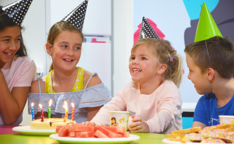 Kids singing happy birthday next to a cake at a table.